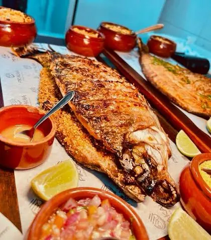 Peixe inteiro assado sobre farofa, com vinagrete, limão e molhos em potes de barro